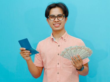 Elinde para tutan gülümseyen bir Asyalı iş adamı ve mavi arka planda izole edilmiş mutlu bir ifadeyi gösteren bir banka tasarruf kitabı..