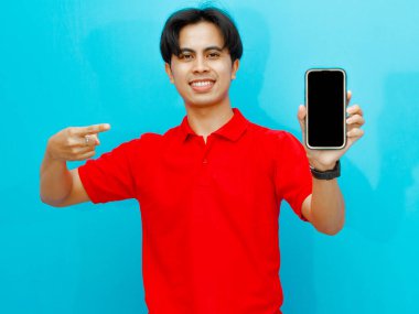 Kırmızı polo tişörtlü Asyalı bir adamın portresi mavi arka planda izole edilmiş mutlu bir ifadeyi gösteren boş bir cep telefonu ekranını işaret ediyor..