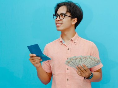 Portrait of a smiling Asian man wearing a pink shirt holding a bank savings book and money, looking to the upper right side with an enthusiastic expression, isolated on a blue background. clipart