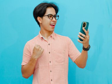 Neşeli bir ifadeyle başarısını kutlarken cep telefonu tutan mutlu Asyalı işadamı. Mavi arka planda izole edilmiş. İş, teknoloji ve yaşam tarzı kavramları için mükemmel..