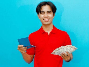 Kırmızı polo tişörtlü Asyalı bir adamın portresi. Elinde para ve banka tasarruf kitabı var. Mavi arka planda gülümseyen mutlu bir ifade var..