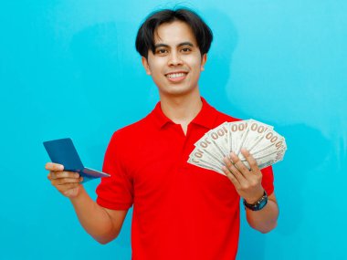 Kırmızı polo tişörtlü Asyalı bir adamın portresi. Elinde para ve banka tasarruf kitabı var. Mavi arka planda gülümseyen mutlu bir ifade var..