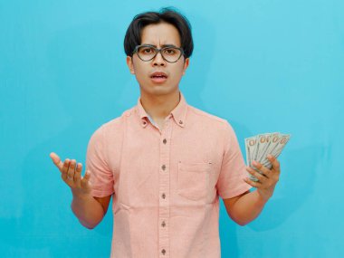 Confused Asian businessman holding money, showing a shocked expression Isolated on blue background. clipart