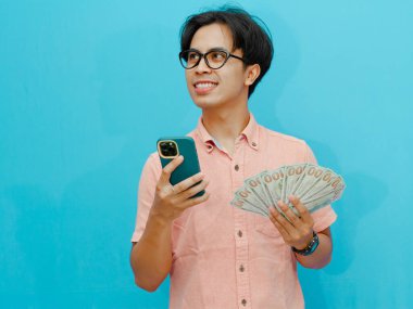 Gülümseyen Asyalı işadamı elinde para ve cep telefonu tutuyor, yukarı bakıyor ve düşünüyor, bir şeyler planlıyor. Mavi arka planda izole edilmiş, iş veya finansal konsept görüntüleri için mükemmel.
