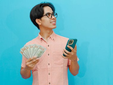Gülümseyen Asyalı işadamı elinde para ve cep telefonu tutuyor, yukarı bakıyor ve düşünüyor, bir şeyler planlıyor. Mavi arka planda izole edilmiş, iş veya finansal konsept görüntüleri için mükemmel.