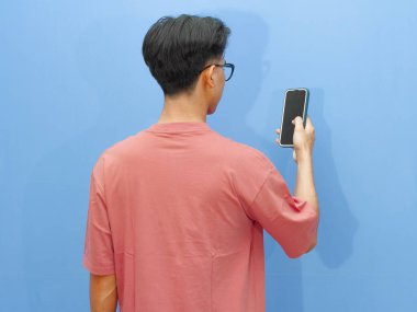 Pembe tişörtlü Asyalı adam mavi arka planda cep telefonu tutarak poz veriyor..