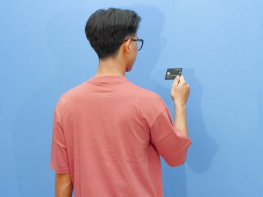 Pembe tişörtlü Asyalı adam mavi arka planda banka kartı tutarak poz veriyor..