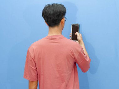 Pembe tişörtlü Asyalı adam mavi arka planda cep telefonu tutarak poz veriyor..