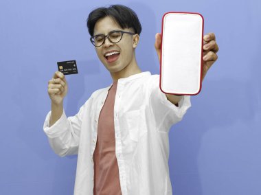 Asyalı bir adam kredi kartı tutuyor ve mavi arka planda heyecanlı, mutlu bir ifadeyle boş bir cep telefonu ekranı gösteriyor..