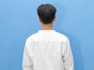 Sıradan giyinmiş Asyalı bir adamın mavi arka planda dikilirken görüntüsü..