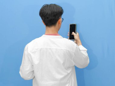 Sıradan giyinmiş Asyalı bir adamın arka planda mavi bir arka planda cep telefonu tutuşu..