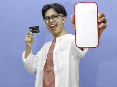 Asyalı bir adam kredi kartı tutuyor ve mavi arka planda heyecanlı, mutlu bir ifadeyle boş bir cep telefonu ekranı gösteriyor..