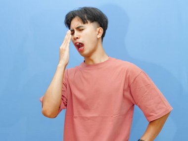 Pembe tişörtlü genç bir adam elini ağzının üstüne koyup mavi arka plana yaslanır..