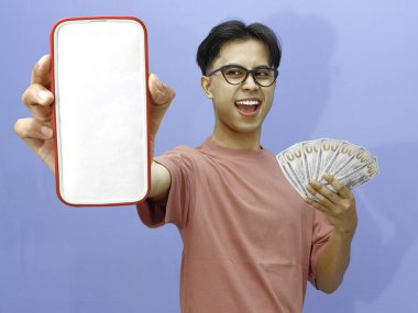 Pembe tişörtlü heyecanlı Asyalı adam elinde mavi arka planda nakit tutarken boş cep telefonu gösteriyor.