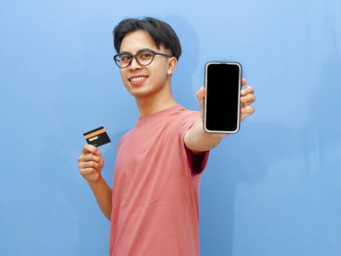 Pembe tişörtlü gülümseyen Asyalı adam elinde bir banka kartı tutarken boş cep telefonu ekranı gösteriyor, mavi arka planda izole edilmiş, reklamlar için mükemmel..