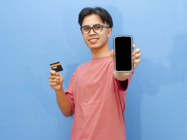 Pembe tişörtlü gülümseyen Asyalı adam elinde bir banka kartı tutarken boş cep telefonu ekranı gösteriyor, mavi arka planda izole edilmiş, reklamlar için mükemmel..