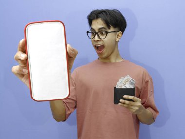 Şaşırmış ifadeye sahip heyecanlı Asyalı adam mavi arka planda içi para dolu boş telefon ekranı ve cüzdan gösteriyor..