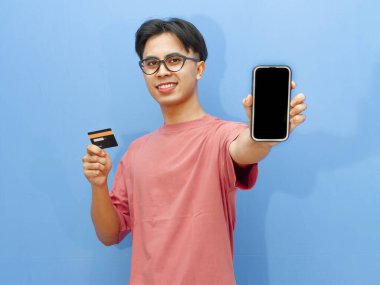 Pembe tişörtlü gülümseyen Asyalı adam elinde bir banka kartı tutarken boş cep telefonu ekranı gösteriyor, mavi arka planda izole edilmiş, reklamlar için mükemmel..