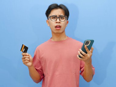 Pembe tişörtlü mutsuz bir Asyalı adamın portresi. Elinde kredi kartı ve cep telefonu var. Şok olmuş bir ifade var..