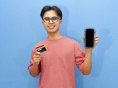 Pembe tişörtlü gülümseyen Asyalı adam elinde bir banka kartı tutarken boş cep telefonu ekranı gösteriyor, mavi arka planda izole edilmiş, reklamlar için mükemmel..