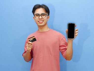 Pembe tişörtlü gülümseyen Asyalı adam elinde bir banka kartı tutarken boş cep telefonu ekranı gösteriyor, mavi arka planda izole edilmiş, reklamlar için mükemmel..