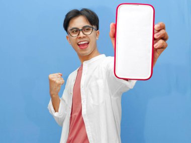 Mutlu Asyalı bir adamın portresi boş bir cep telefonu ekranını tutuyor, ellerini kaldırıyor ve başarıyı mavi arka planda heyecanlı bir ifadeyle kutluyor..