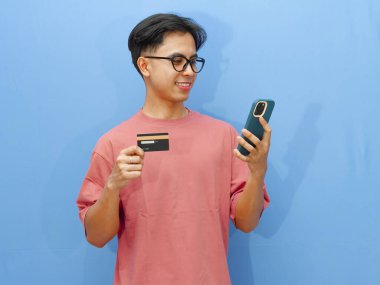 Pembe tişörtlü heyecanlı Asyalı adam elinde bir kredi kartı ve mutlu bir ifadeyle akıllı telefon kullanıyor, e-ticaret ya da reklam için mükemmel, mavi arka planda izole edilmiş..