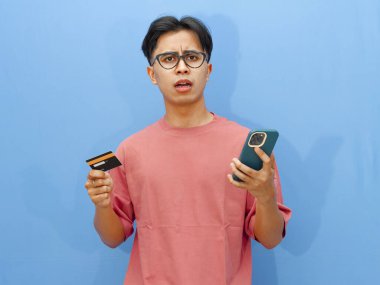 Pembe tişörtlü mutsuz bir Asyalı adamın portresi. Elinde kredi kartı ve cep telefonu var. Şok olmuş bir ifade var..
