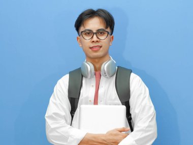 Portrait of a happy Asian male college student wearing a casual wear, holding a laptop, carrying backpack and smiling confidently while posing on an isolated blue background. clipart