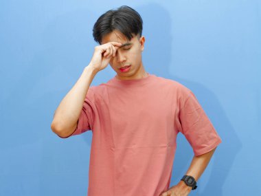 Sağlıksız Asyalı bir adam üzgün bir ifadeyle başını tutup mavi bir arka planda tek başına duruyor..