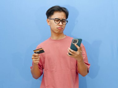 Pembe tişörtlü mutsuz bir Asyalı adamın portresi. Elinde kredi kartı ve cep telefonu var. Şok olmuş bir ifade var..