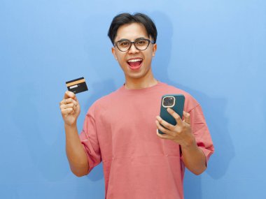 Pembe tişörtlü heyecanlı Asyalı adam elinde bir kredi kartı ve mutlu bir ifadeyle akıllı telefon kullanıyor, e-ticaret ya da reklam için mükemmel, mavi arka planda izole edilmiş..