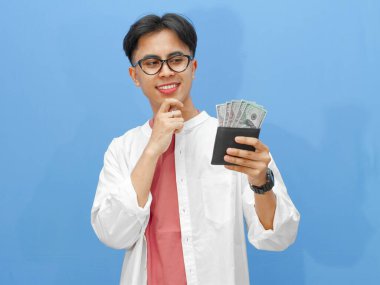 Sıradan giysiler içinde Asyalı bir adamın portresi. Elinde para dolu bir cüzdan, mavi arka planda bir düşünce jesti ile yukarı bakıyor..