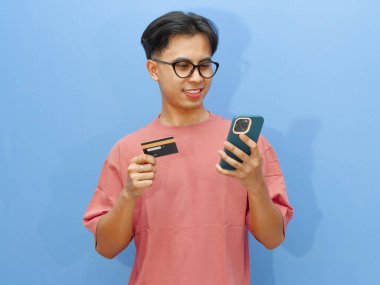 Elinde kredi kartı ve cep telefonu olan gülümseyen Asyalı bir adamın portresi, mavi arka planda izole edilmiş bir şekilde online ödeme yapıyor..