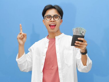 Mavi arka planda soyutlanmış, içi para dolu bir cüzdan taşıyan heyecanlı Asyalı genç bir adam. Reklam, promosyon ya da finansal temalar için mükemmel..