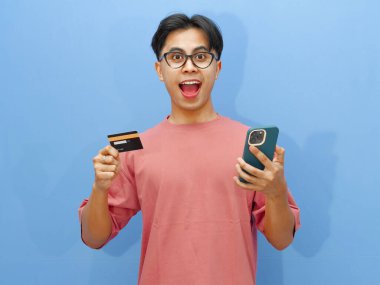 Kredi kartı tutan Asyalı bir adamın portresi ve çevrimiçi ödeme için cep telefonu kullanımı, mavi bir arka planda izole edilmiş, komik ve şaşırtıcı bir ifadeyi gösteriyor..