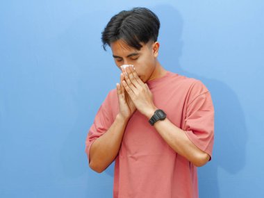 Hasta bir genç adam yüzüne bir mendil dayayıp burnunu acı dolu bir ifadeyle silmeye çalışmış, mavi bir arka planda izole edilmiş..