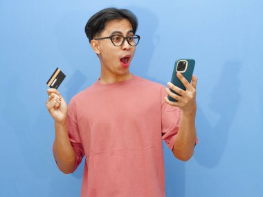 Kredi kartı tutan Asyalı bir adamın portresi ve çevrimiçi ödeme için cep telefonu kullanımı, mavi bir arka planda izole edilmiş, komik ve şaşırtıcı bir ifadeyi gösteriyor..