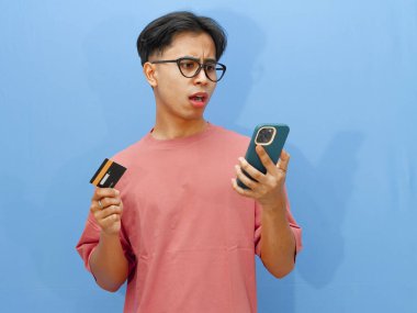 Pembe tişörtlü mutsuz bir Asyalı adamın portresi. Elinde kredi kartı ve cep telefonu var. Şok olmuş bir ifade var..