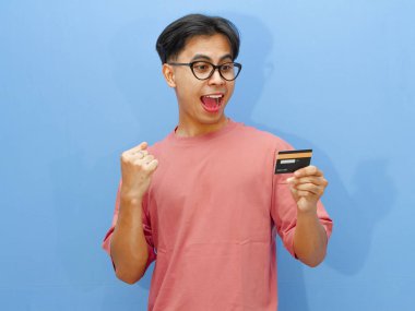 Elinde kredi kartı olan heyecanlı bir Asyalı adamın portresi zaferi bir çığlık ve mavi arka planda sıkılmış yumruklarla kutluyor..
