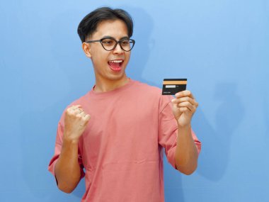Elinde kredi kartı olan heyecanlı bir Asyalı adamın portresi zaferi bir çığlık ve mavi arka planda sıkılmış yumruklarla kutluyor..