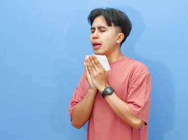 Elinde doku tutan sağlıksız genç adam hapşırmaya hazırlanıyor. Üzüntü ve acıyı ifade ediyor. Mavi arka planda izole edilmiş..