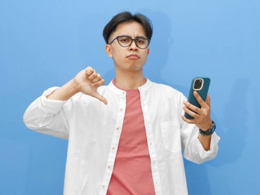 Cep telefonu tutan Asyalı bir adamın portresi, mavi arka planda hayal kırıklığına uğramış bir ifadeyle baş parmağını kaldırıyor..