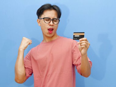 Elinde kredi kartı olan heyecanlı bir Asyalı adamın portresi zaferi bir çığlık ve mavi arka planda sıkılmış yumruklarla kutluyor..