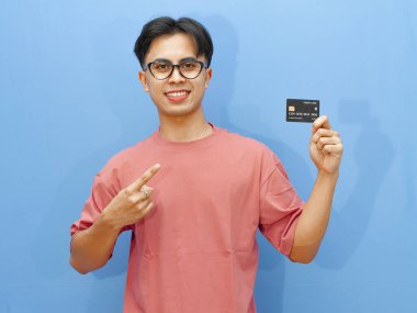 Asyalı adam mavi arka planda mutlu ve heyecanlı bir ifadeyle kredi kartını işaret ediyor. Finansal reklamlar ve terfiler için mükemmel..