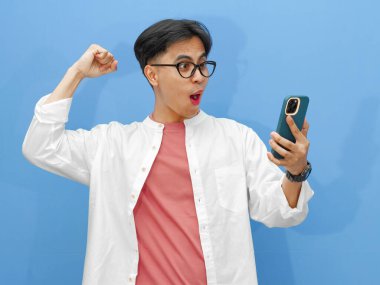 Mutlu Asyalı genç adam, mavi arka planda heyecanla el kaldırarak akıllı telefonuna bakarken başarısını kutluyor..