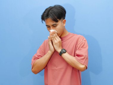 Hasta bir genç adam yüzüne bir mendil dayayıp burnunu acı dolu bir ifadeyle silmeye çalışmış, mavi bir arka planda izole edilmiş..