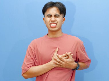 Sağlıksız Asyalı bir adam mavi arka planda acı ve üzüntüyle göğsüne dokunuyor..