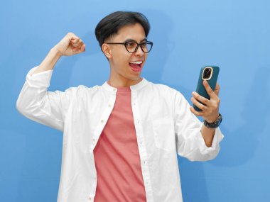 Mutlu Asyalı genç adam, mavi arka planda heyecanla el kaldırarak akıllı telefonuna bakarken başarısını kutluyor..