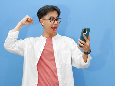 Mutlu Asyalı genç adam, mavi arka planda heyecanla el kaldırarak akıllı telefonuna bakarken başarısını kutluyor..